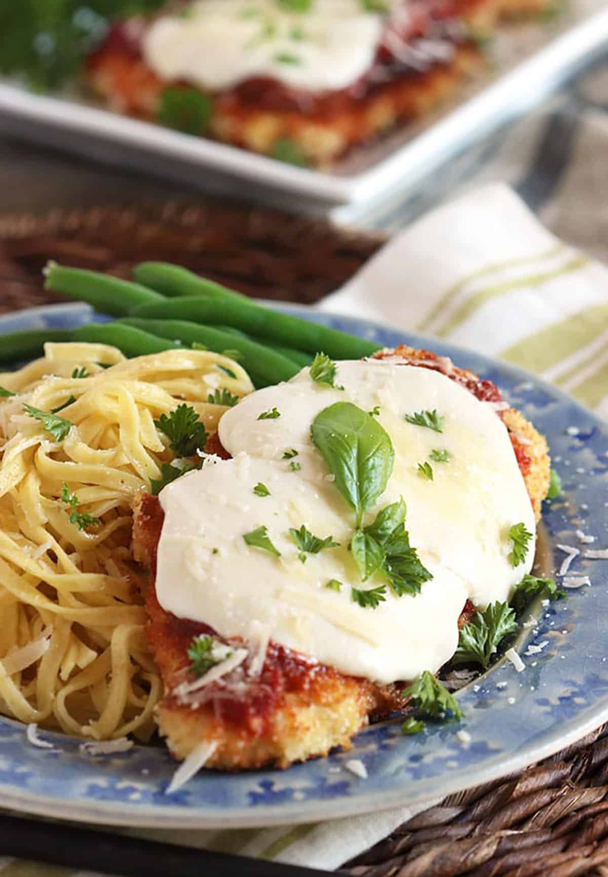 Chicken parmesan topped with basil on a blue plate with pasta and green beans.