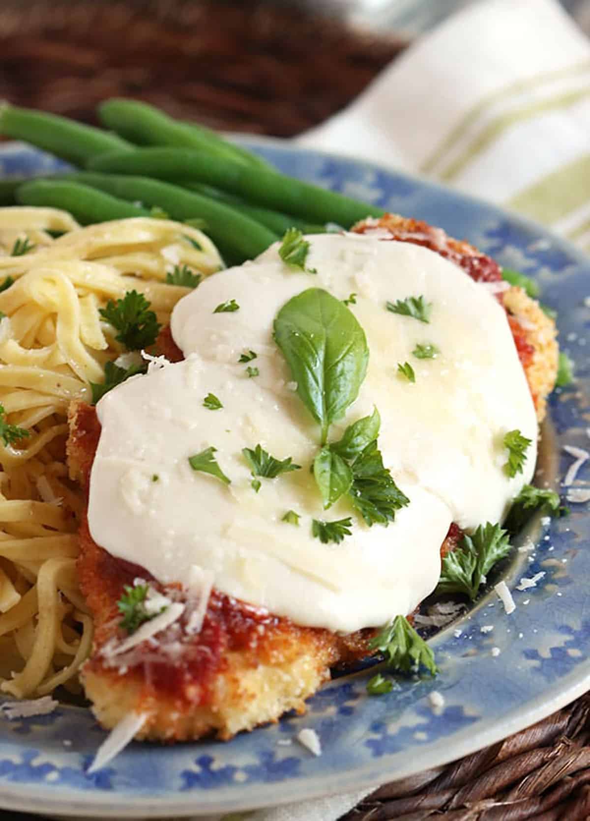 chicken Parmesan on a blue plate topped with a basil sprig and a side of pasta with green beans.
