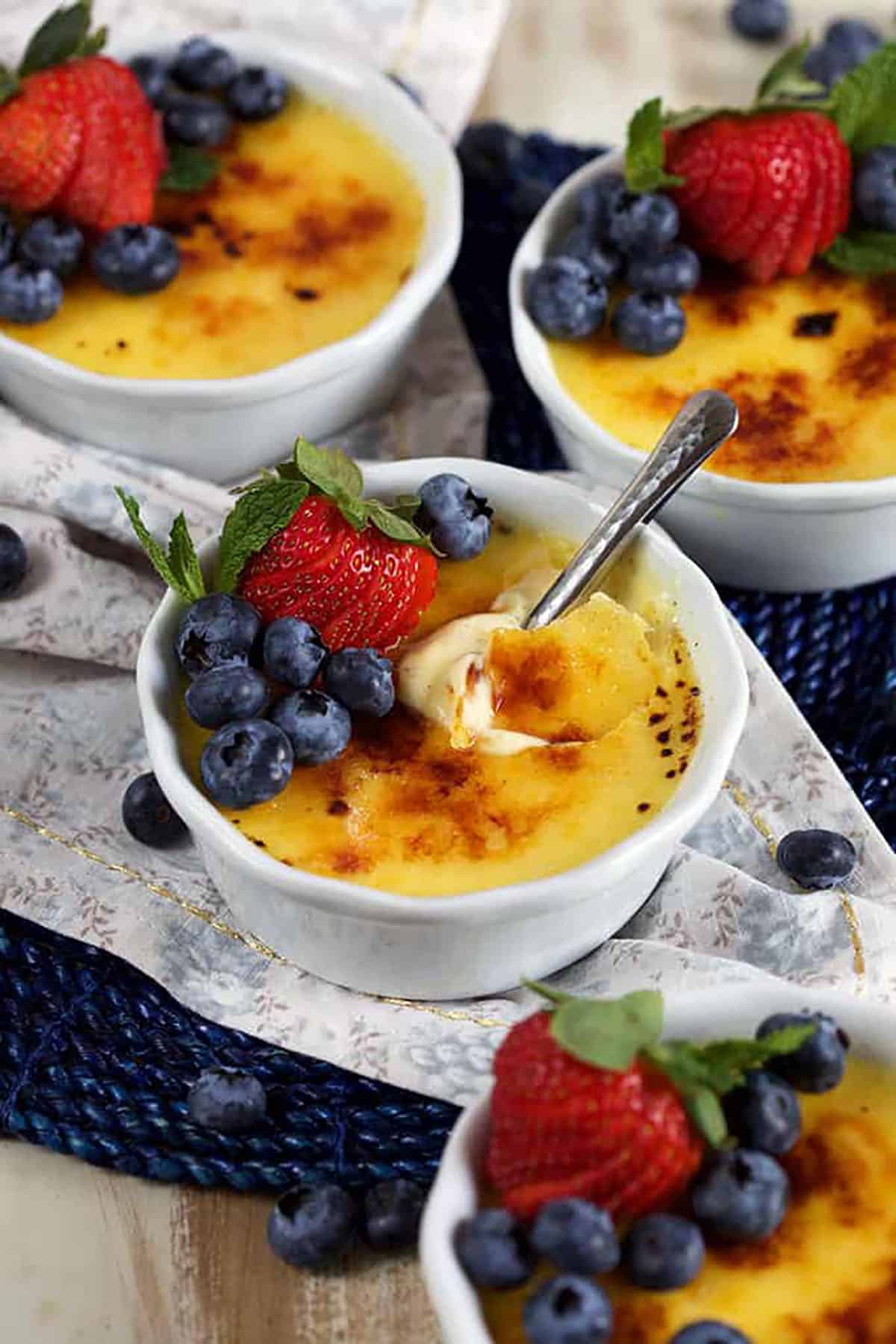 Four white ramekins with creme brûlée topped with berries on a floral napkin
