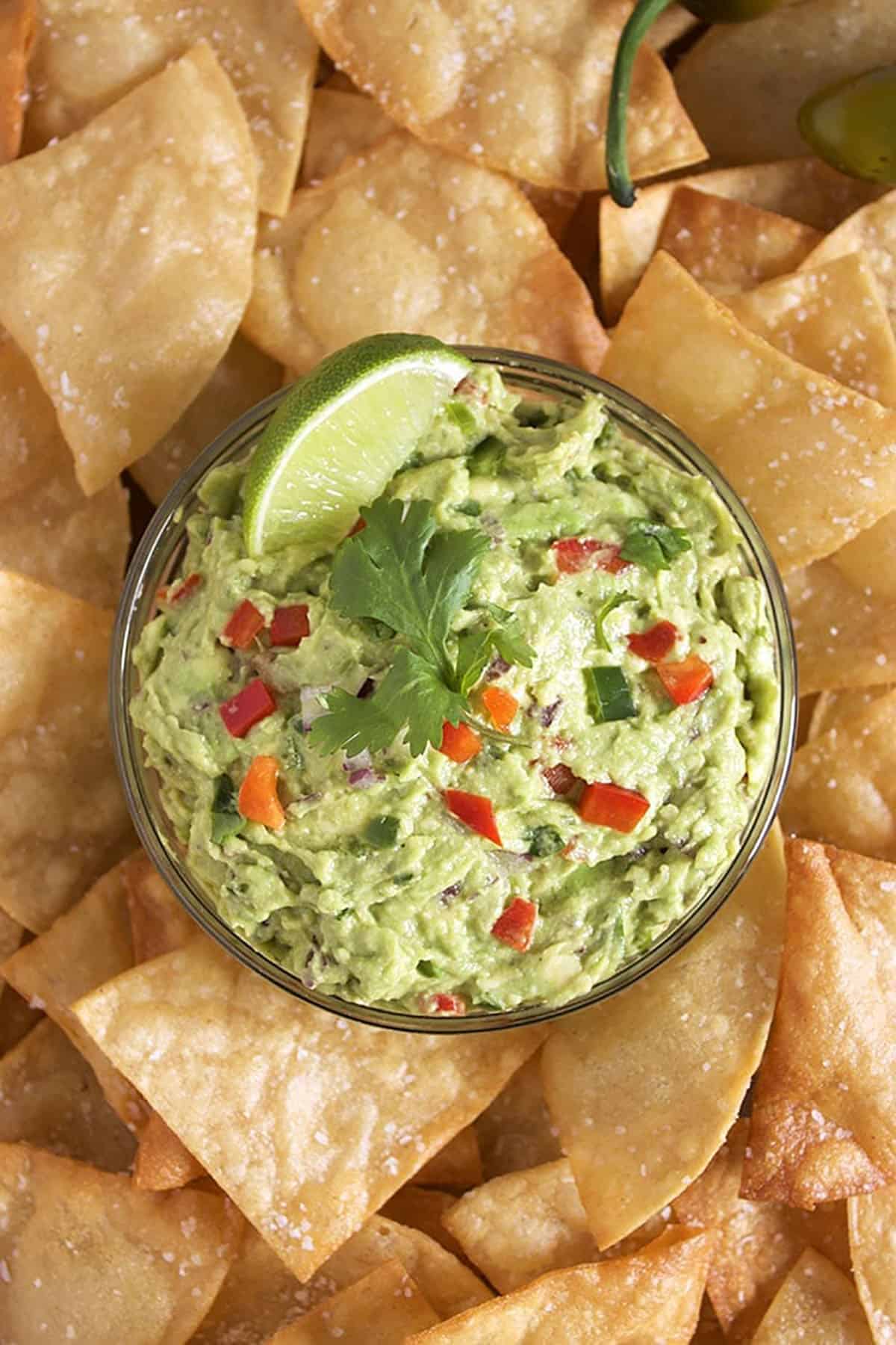 Guacamole in a glass bowl with a lime and cilantro on top surrounded by tortilla chips.