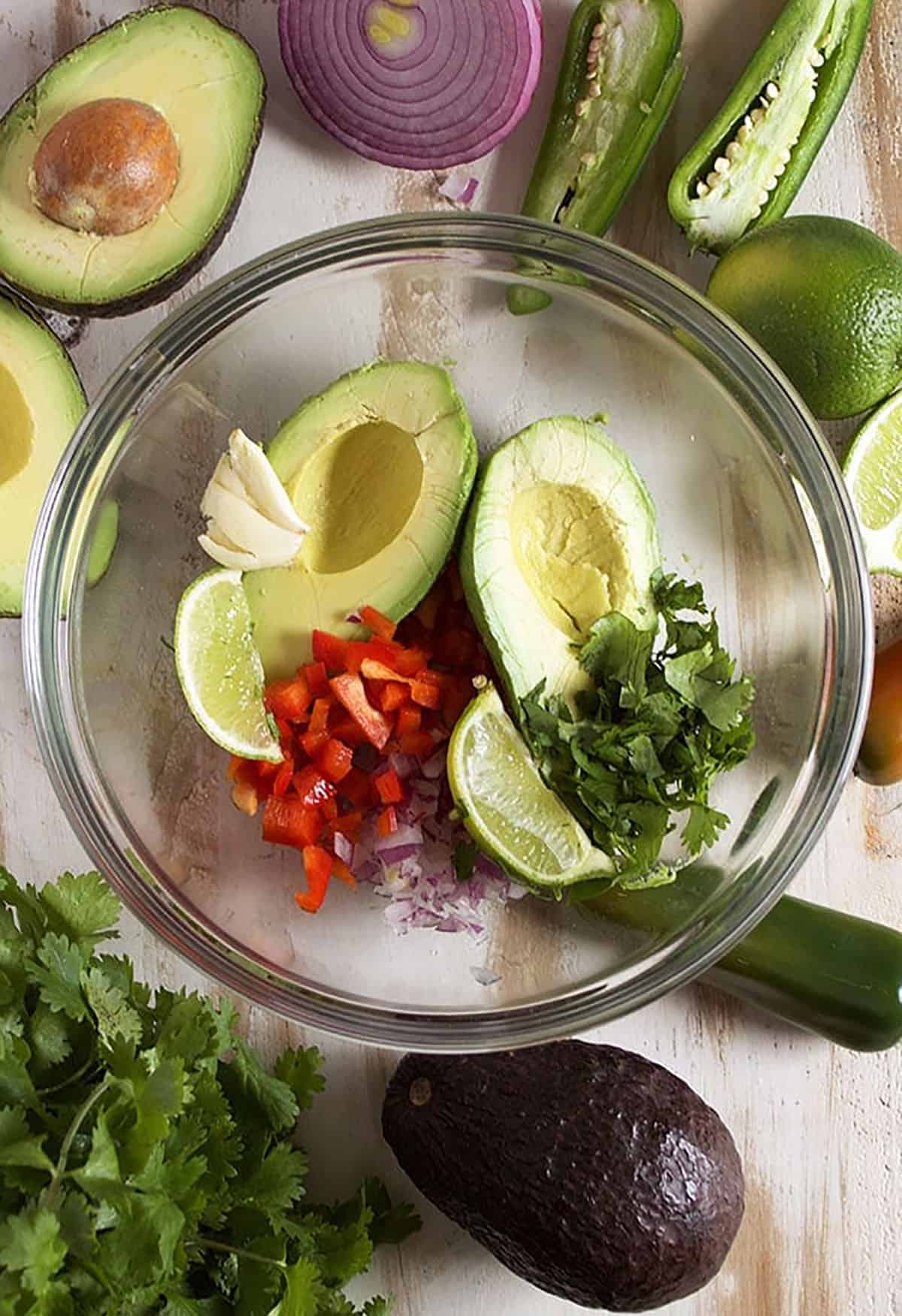 Guacamole ingredients in a glass bowl with half an avocado, jalapeno cut in half and cilantro.