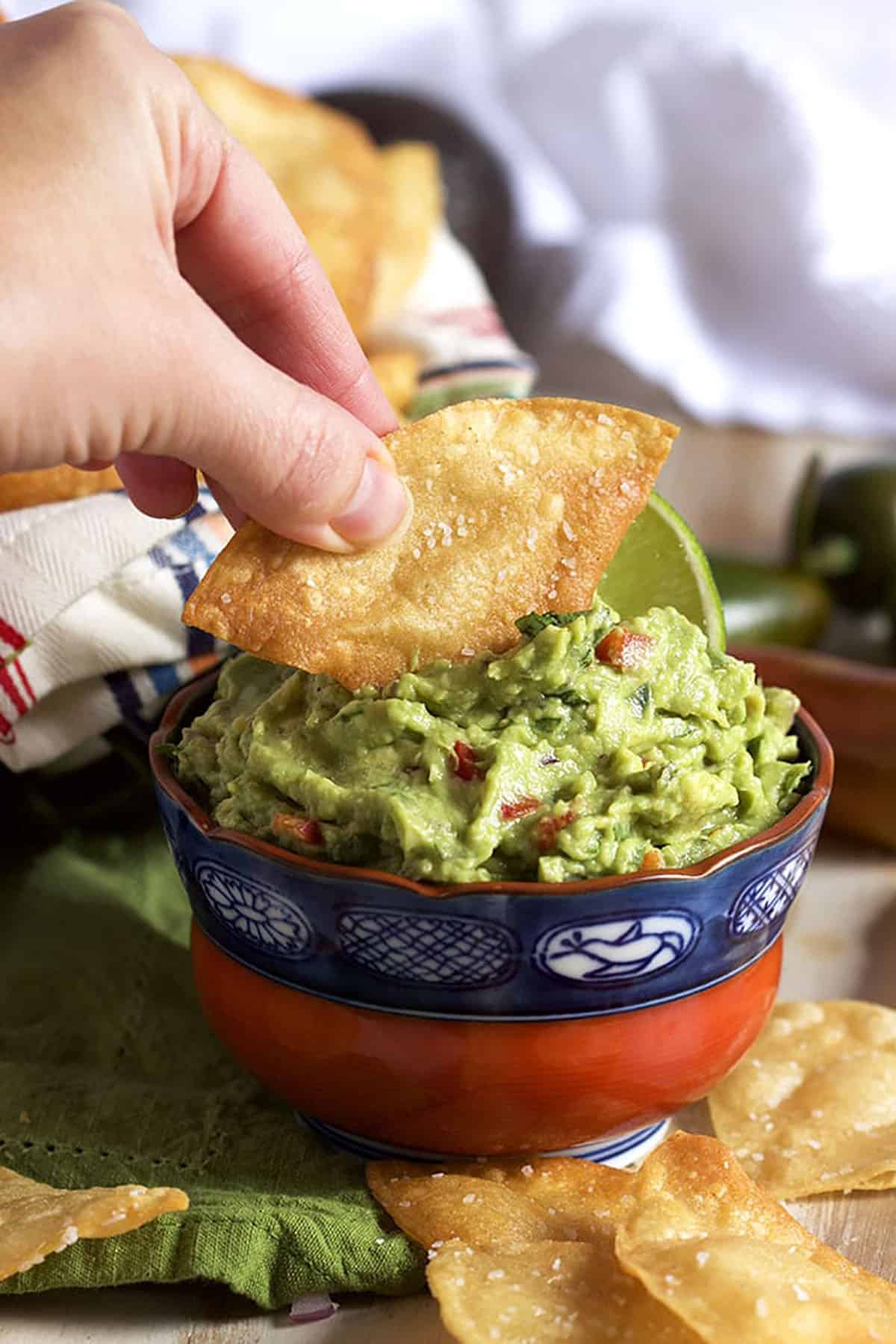 red and blue bowl with guacamole and a hand holding a tortilla chip dipping it into the bowl.