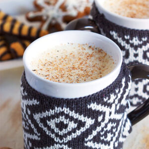 Two mugs with sweaters filled with eggnog with gingerbread cookies in the background.