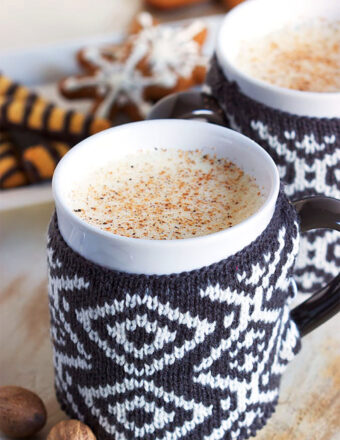 Two mugs with sweaters filled with eggnog with gingerbread cookies in the background.