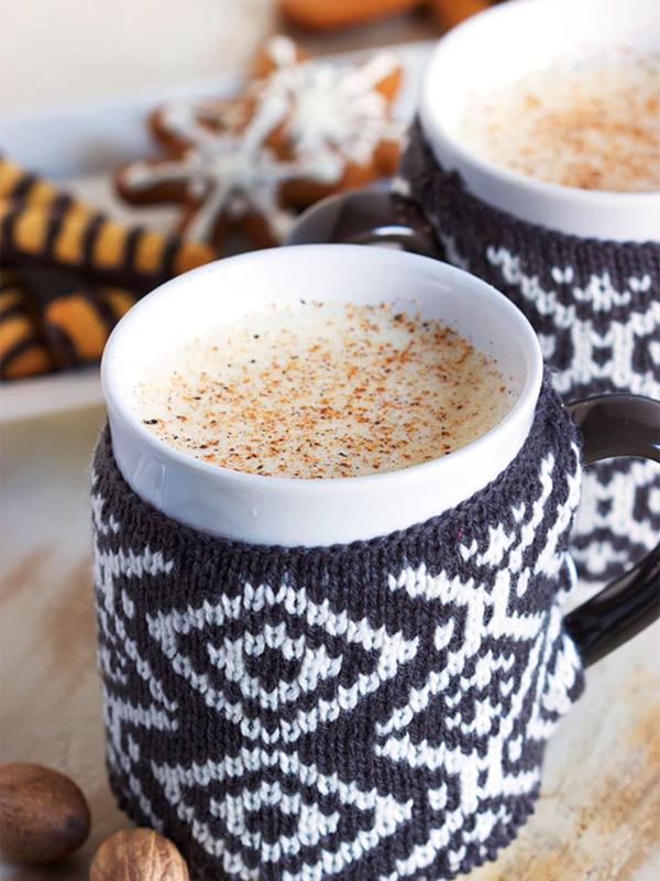 Two mugs with sweaters filled with eggnog with gingerbread cookies in the background.