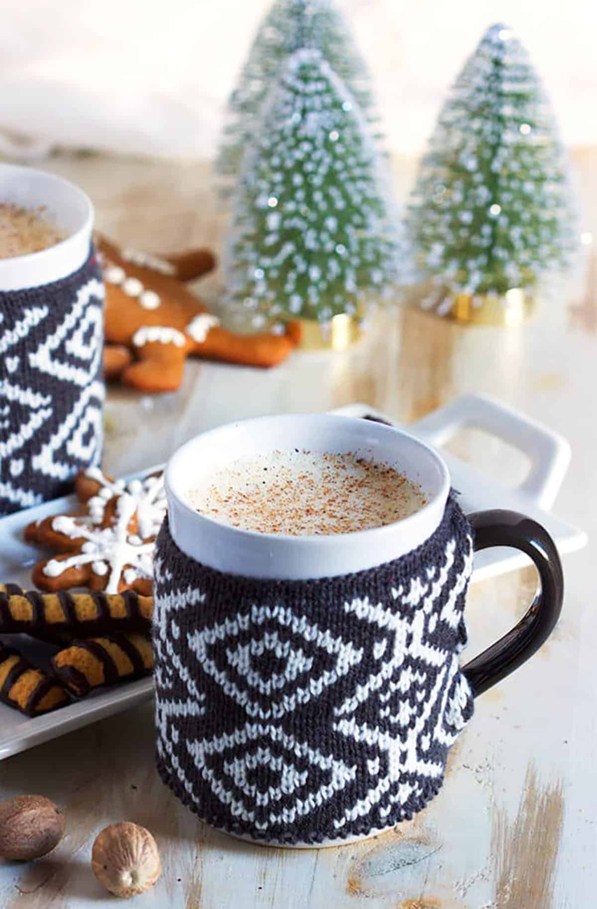 Mug with a knit cozy filled with eggnog with two bottle brush trees in the background.
