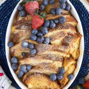An oval shaped white casserole dish is filled with baked french toast casserole and topped with berries and powdered sugar.