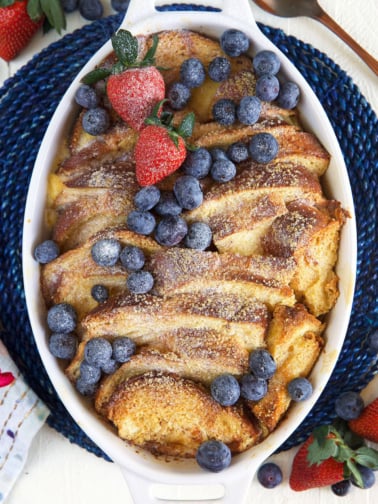 An oval shaped white casserole dish is filled with baked french toast casserole and topped with berries and powdered sugar.
