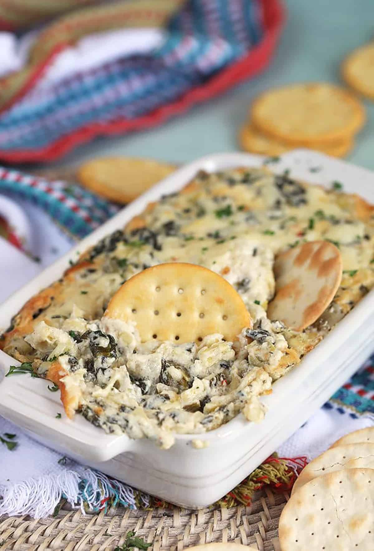 spinach artichoke dip in a white baking dish with a cracker dipped into it.
