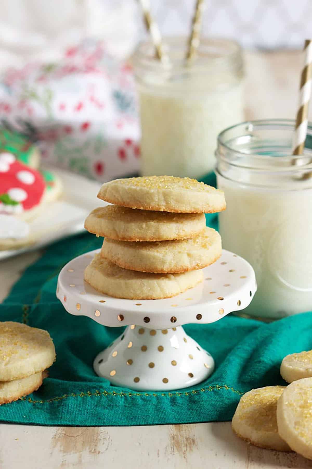 Slice and bake sugar cookies on a cupcake stand placed on a green napkin with glasses of milk in the background. The sugar cookies are stacked on top of each other.