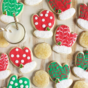 Sugar Cookies shaped like mittens and decorated with red and green royal icing are scattered on a flat wood surface.
