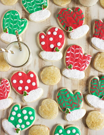 Sugar Cookies shaped like mittens and decorated with red and green royal icing are scattered on a flat wood surface.