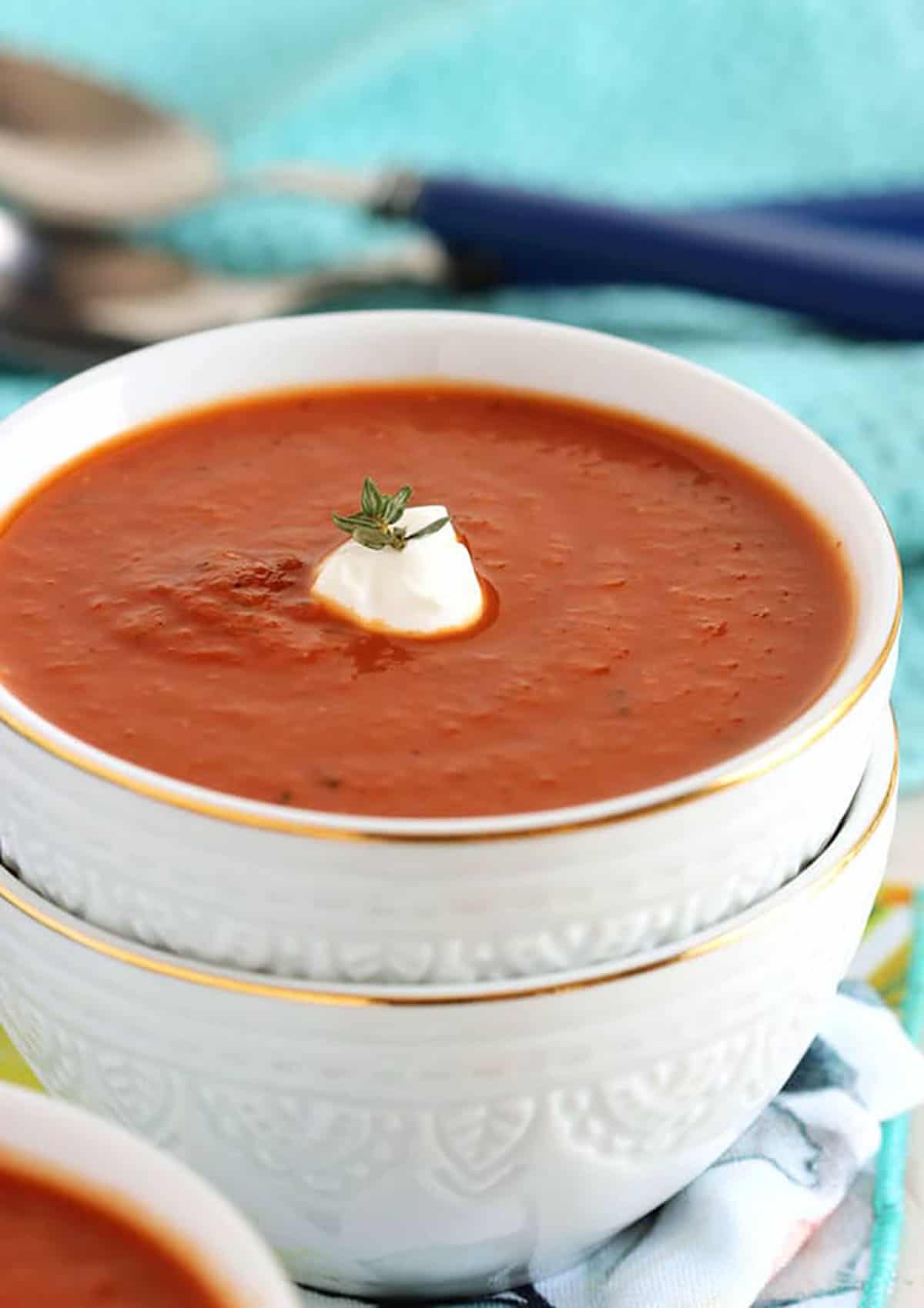 tomato soup with a dollop of sour cream and a sprig of thyme in a white bowl stacked on a another white bowl.