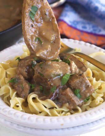 A wooden spoon is drizzling some gravy over a serving of beef tips and noodles.