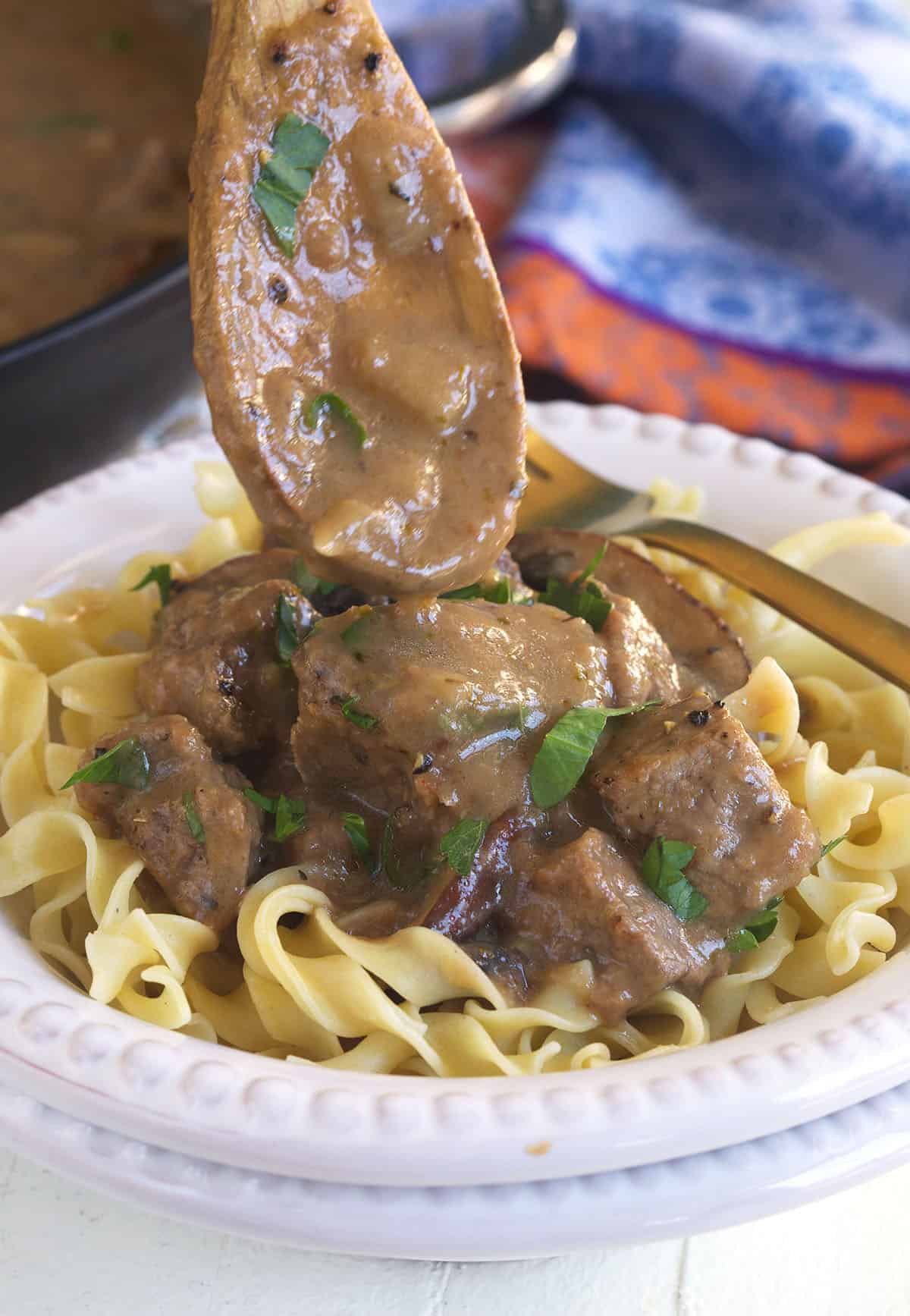 A wooden spoon is drizzling some gravy over a serving of beef tips and noodles.