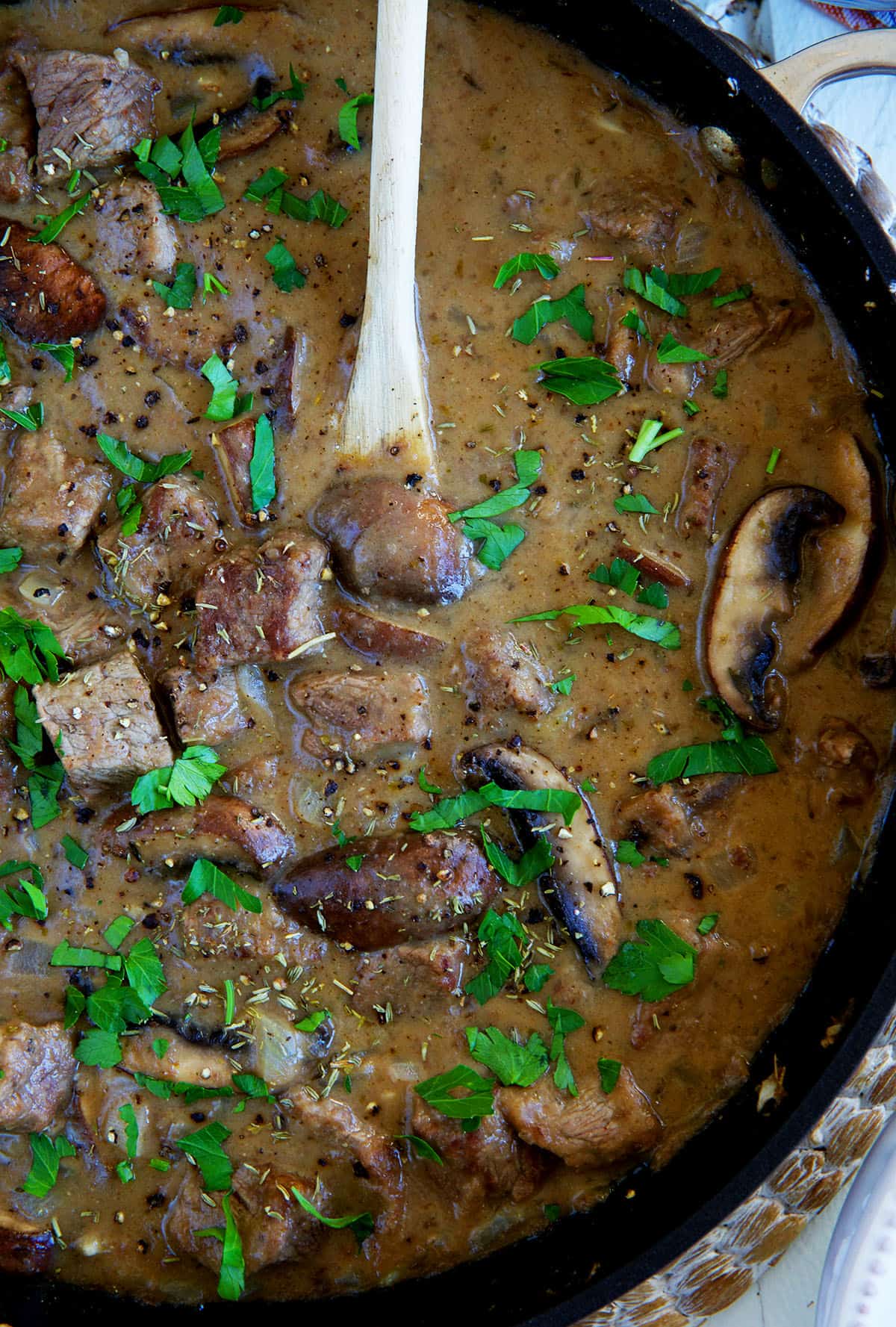 A wooden spoon is stirring beef tips and gravy in a skillet. 