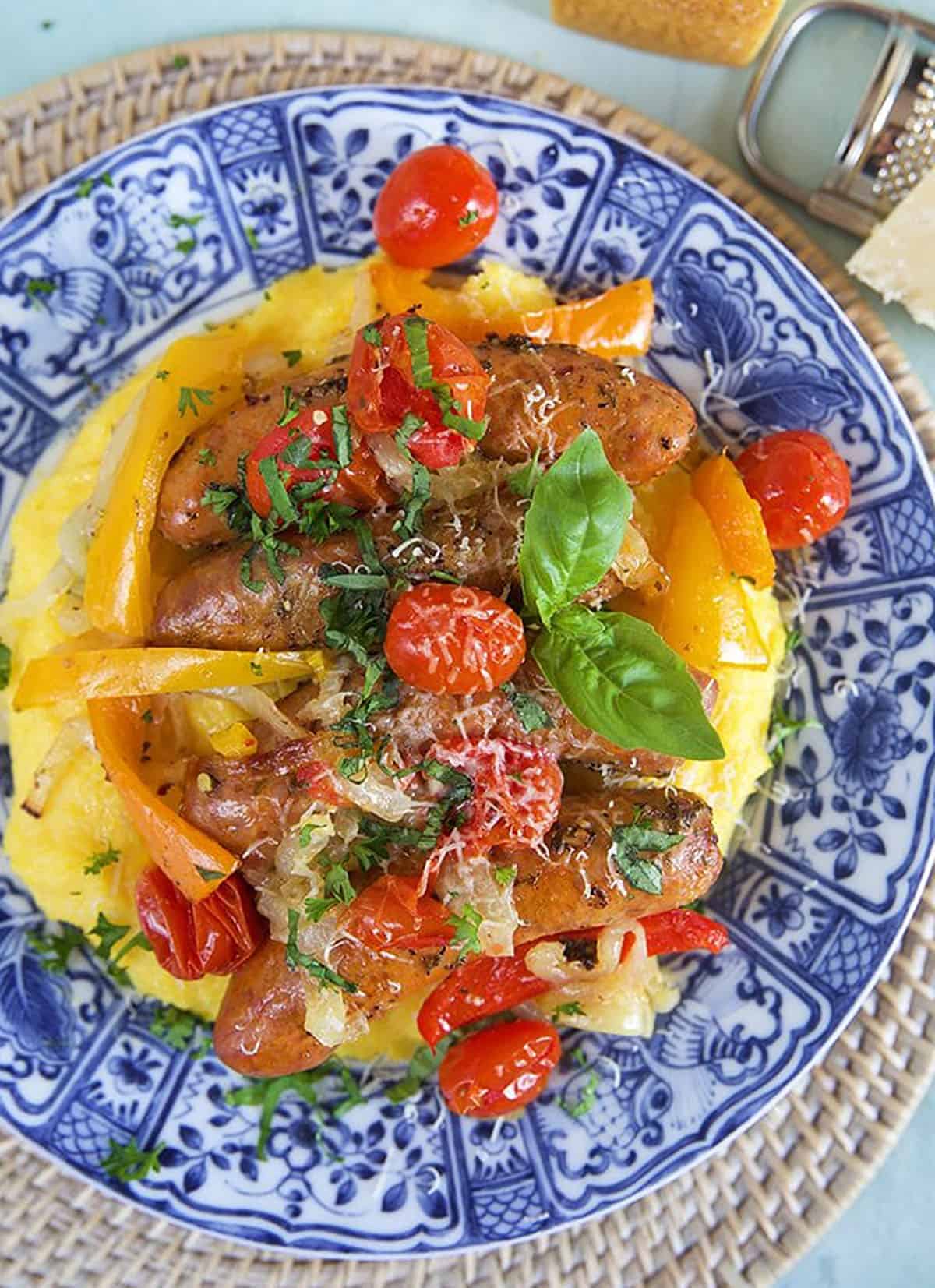 Two links of italian sausage and peppers with onions on a bed of polenta on a blue and white plate.