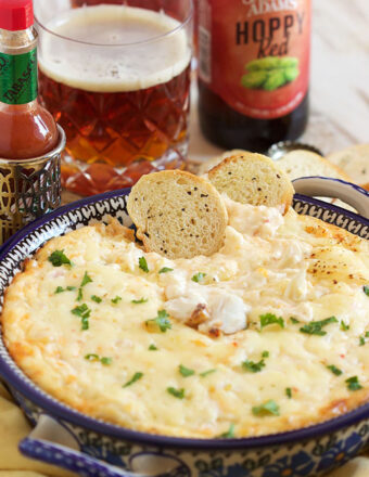 Crab Dip in a polish pottery baking dish with a glass of beer and a bottle of hot sauce in the background.