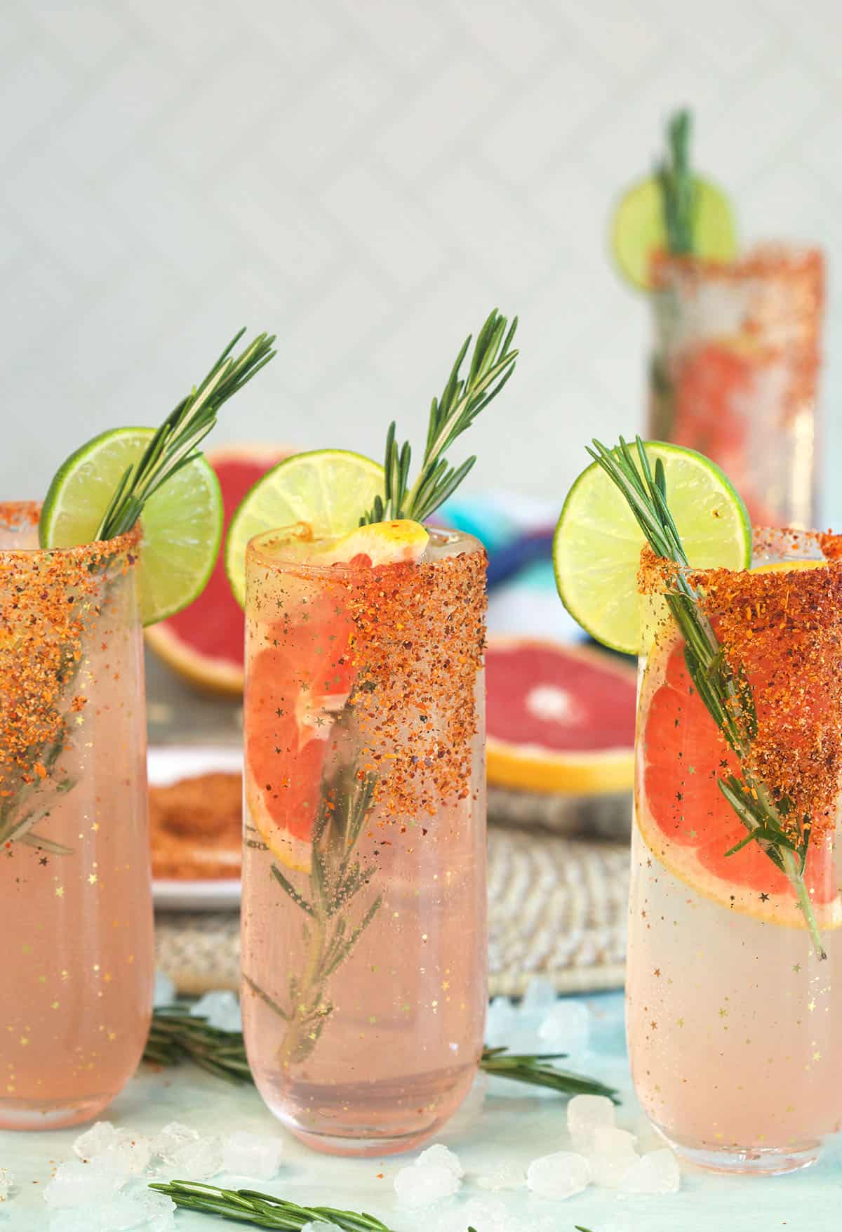 Several glasses filled with paloma cocktail are garnished with rosemary and limes.