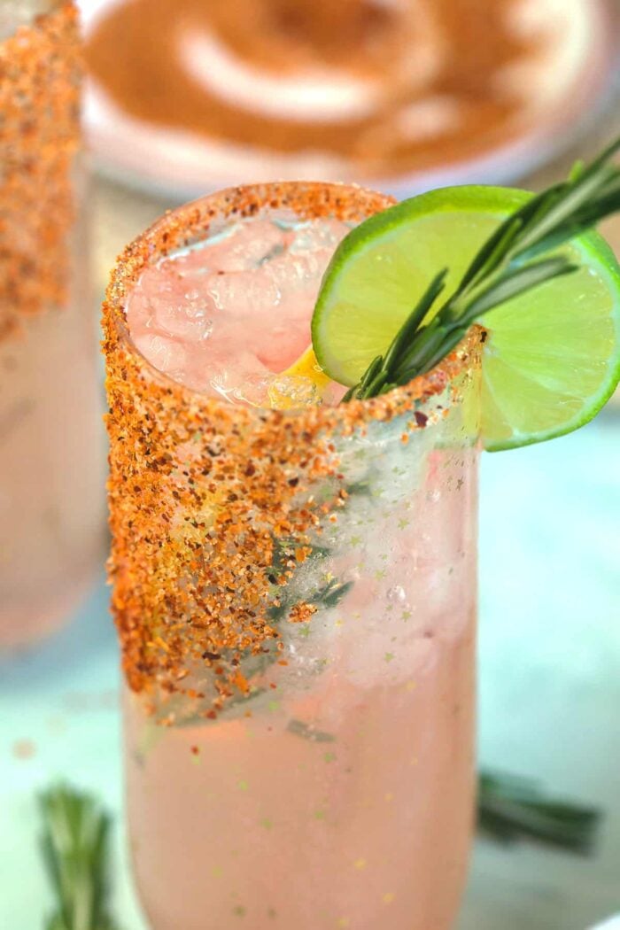 A paloma is garnished with a tajin rim, a lime wedge, and a rosemary sprig.