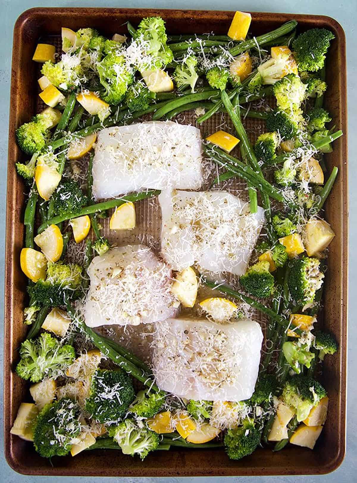 4 cod filets on a baking sheet with veggies for a sheet pan dinner before baking.
