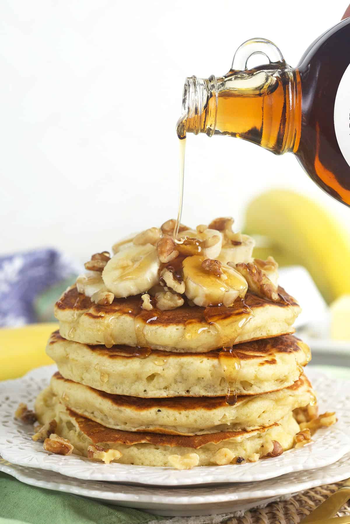 Syrup is being poured all over a stack of garnished banana pancakes.