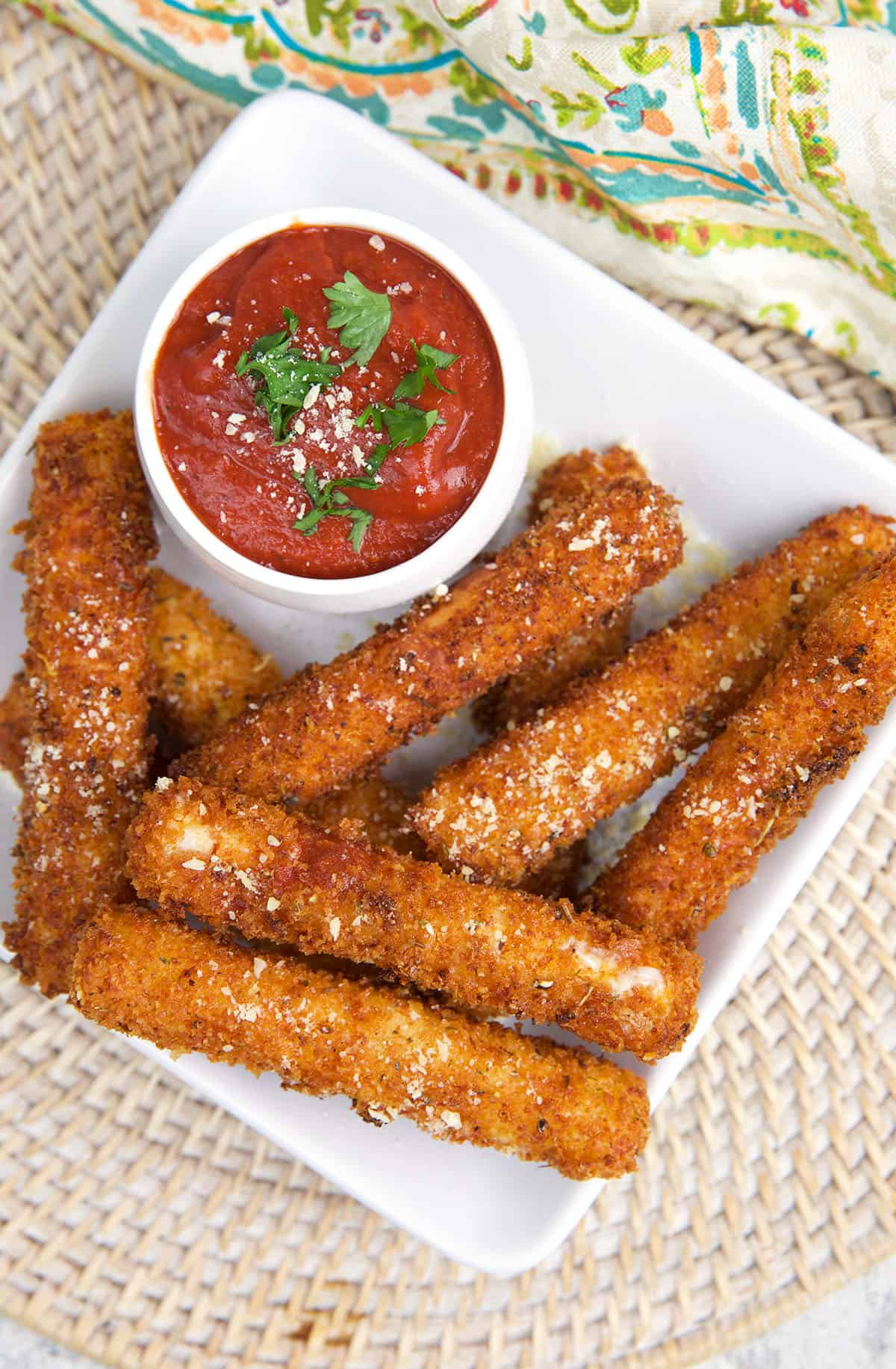 Mozzarella sticks are placed on a white plate next to a small bowl of red sauce.