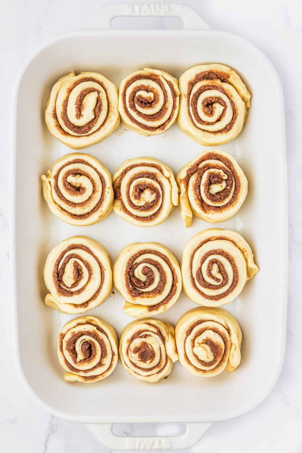 A dozen unbaked cinnamon rolls are placed in a white baking dish.