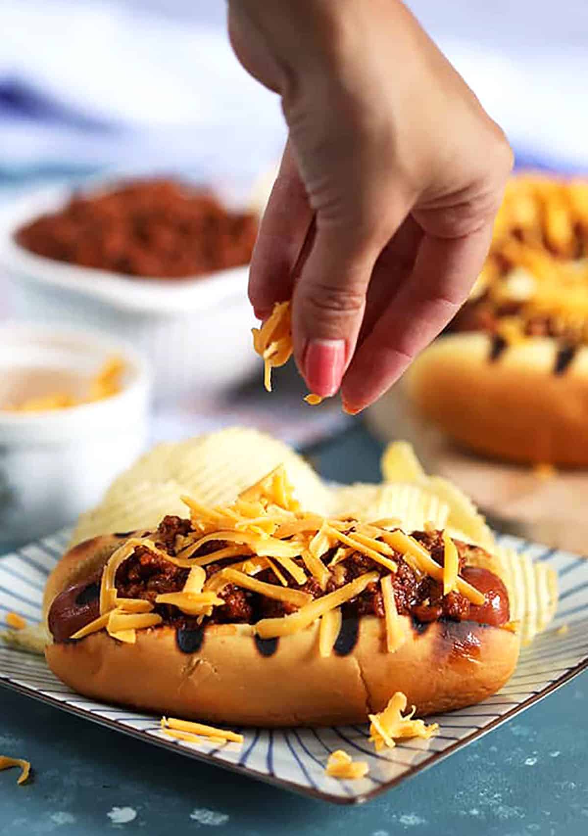 Hand placing shredded cheese on a chili cheese dog