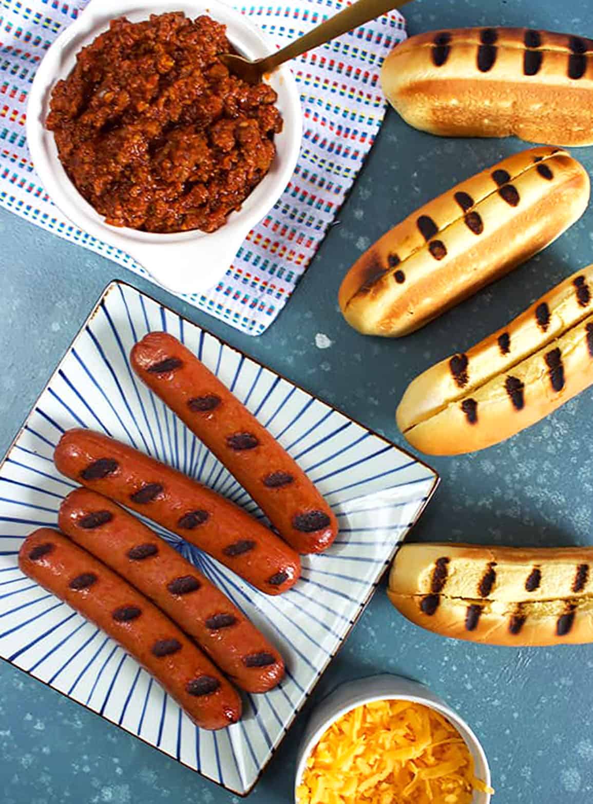 Undressed hot dogs with grilled hot dog rolls, a bowl of chili and shredded cheese