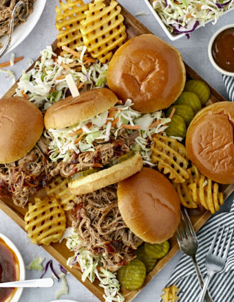 Several pulled pork sandwiches are presented on a serving plate with pickles, fries, and coleslaw.