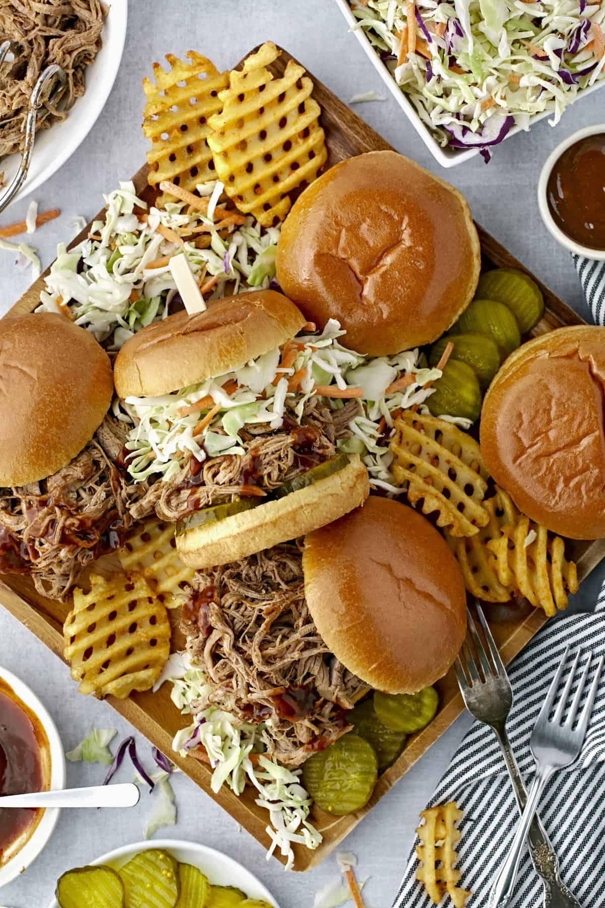Several pulled pork sandwiches are presented on a serving plate with pickles, fries, and coleslaw. 