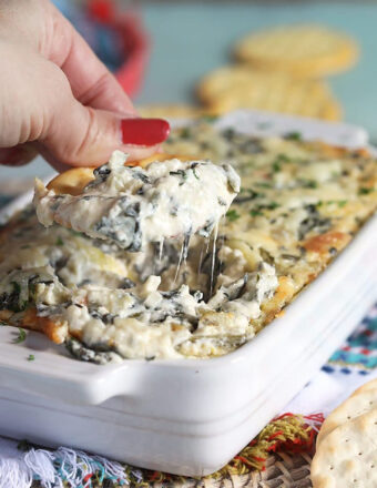 Hand with a cracker taking a dip from Spinach Artichoke Dip baked in a white baking dish.