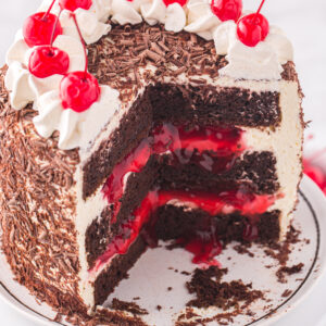 A black forest cake is topped with cherries and whipped cream. The center is cut to reveal a layered inside.