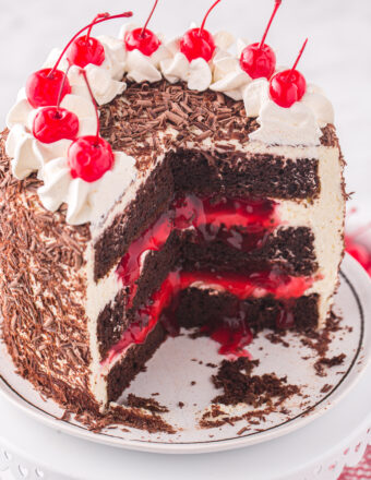 A black forest cake is topped with cherries and whipped cream. The center is cut to reveal a layered inside.