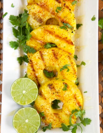 Grilled pineapple on a white platter with sliced lime and chopped cilantro