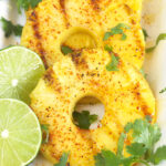 Three slices of grilled pineapple with cilantro sprigs and two lime slices.
