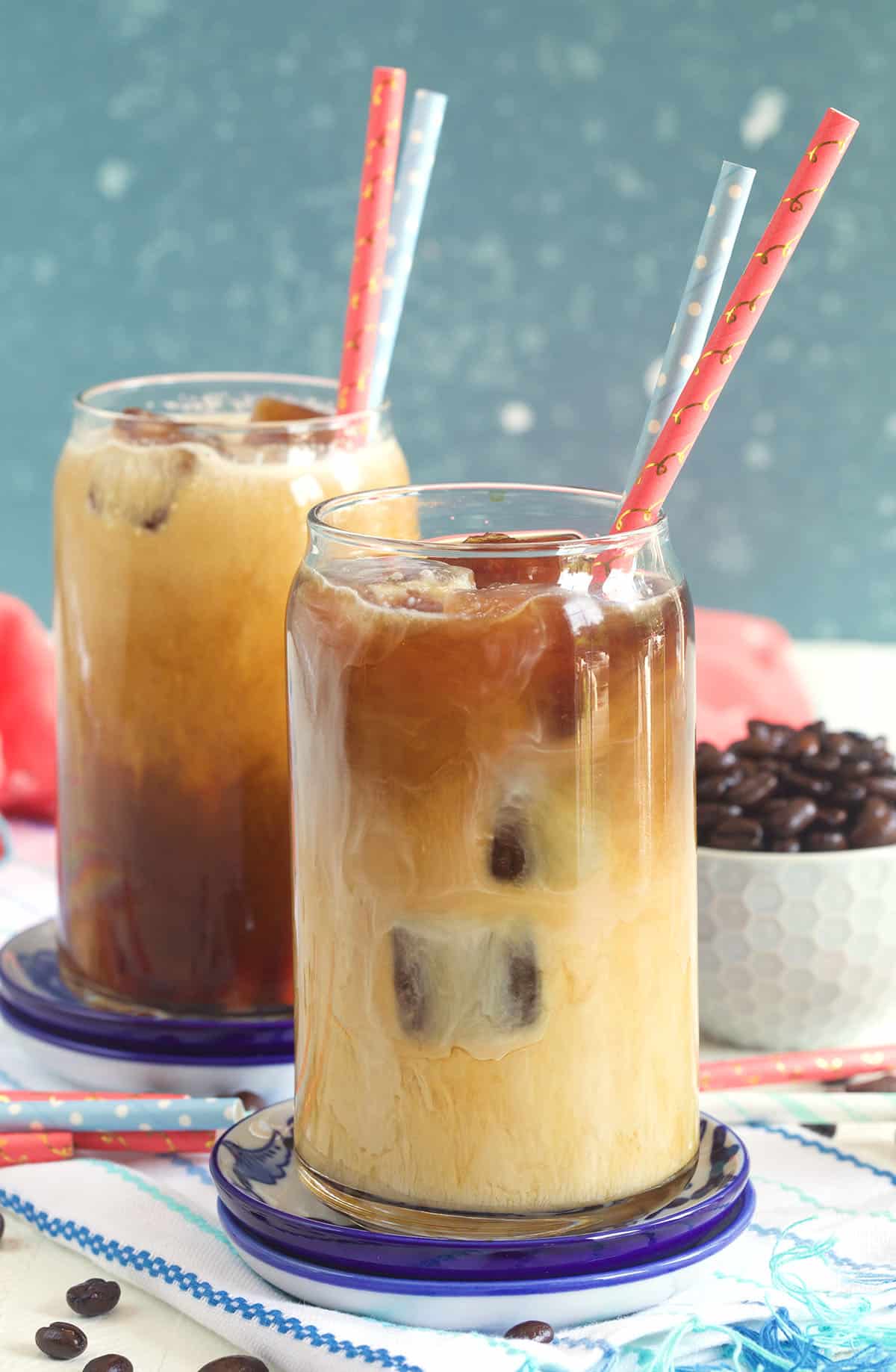 Iced coffee in a glass with coffee ice cubes and two paper straws with a bowl of coffee beans behind it.