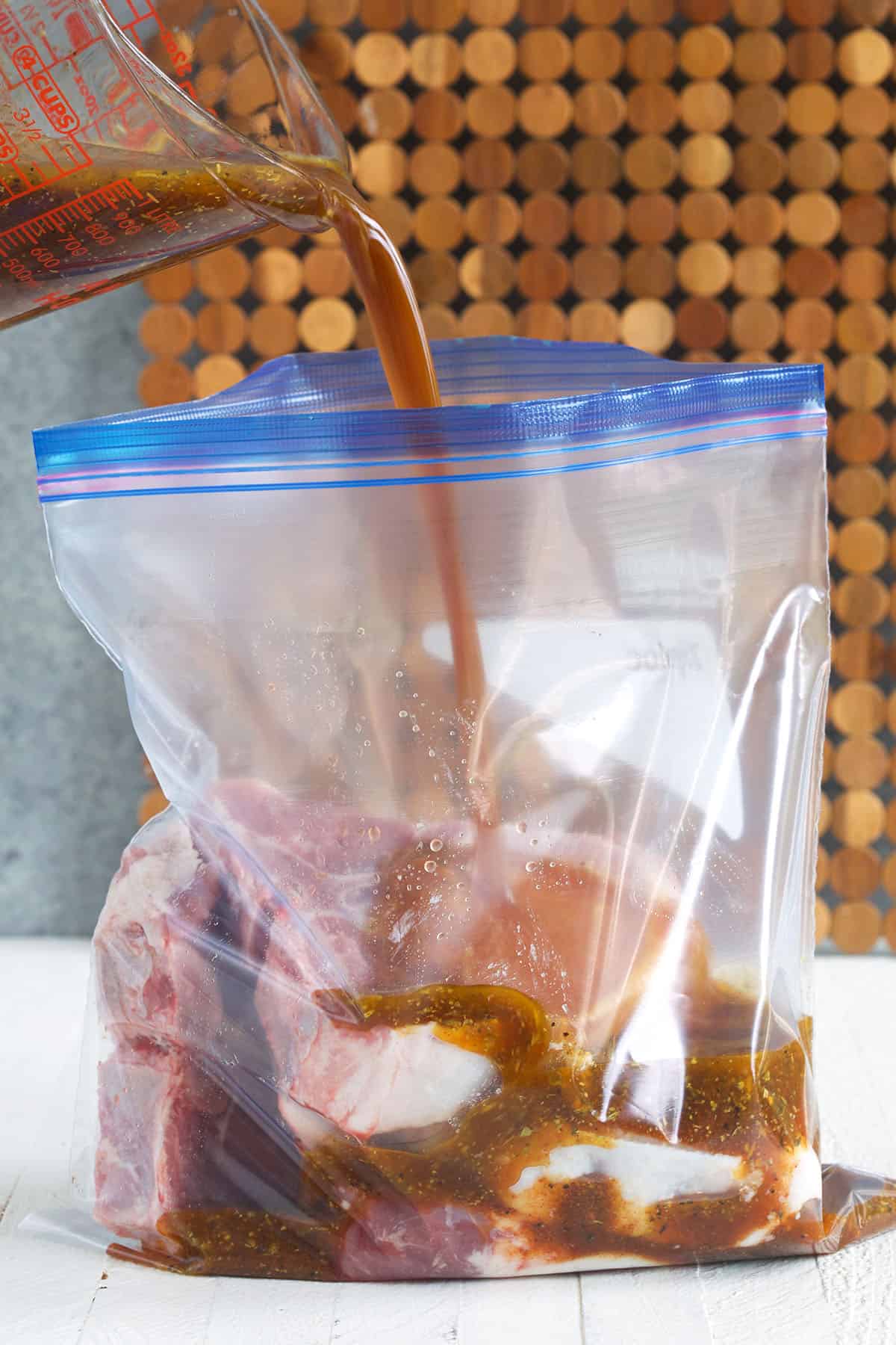 Marinade is being poured into a large zip-top bag filled with raw pork chops. 