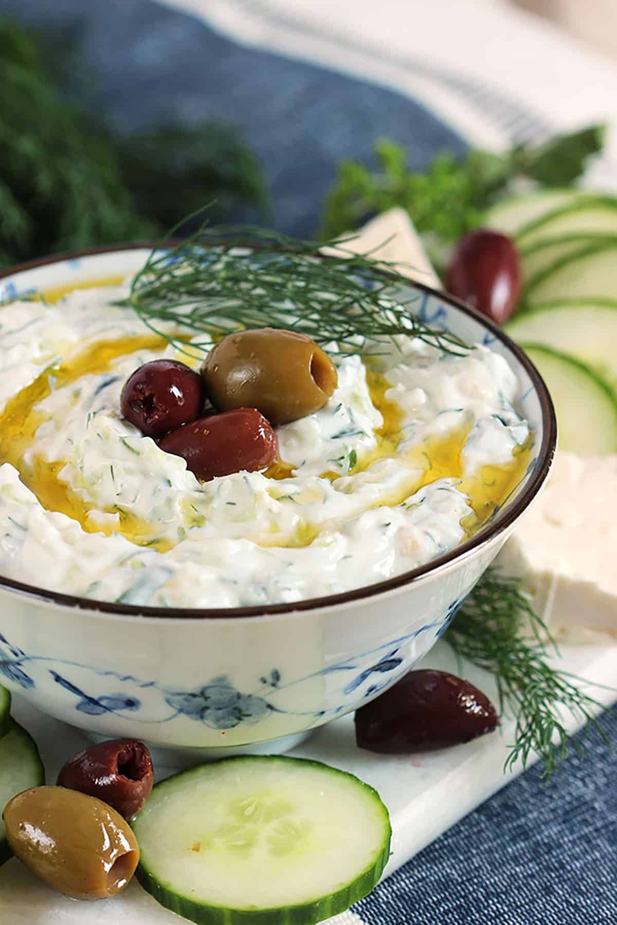 Tzatziki Sauce in a blue and white bowl with kalamata olives on top and olive oil drizzles around it. 