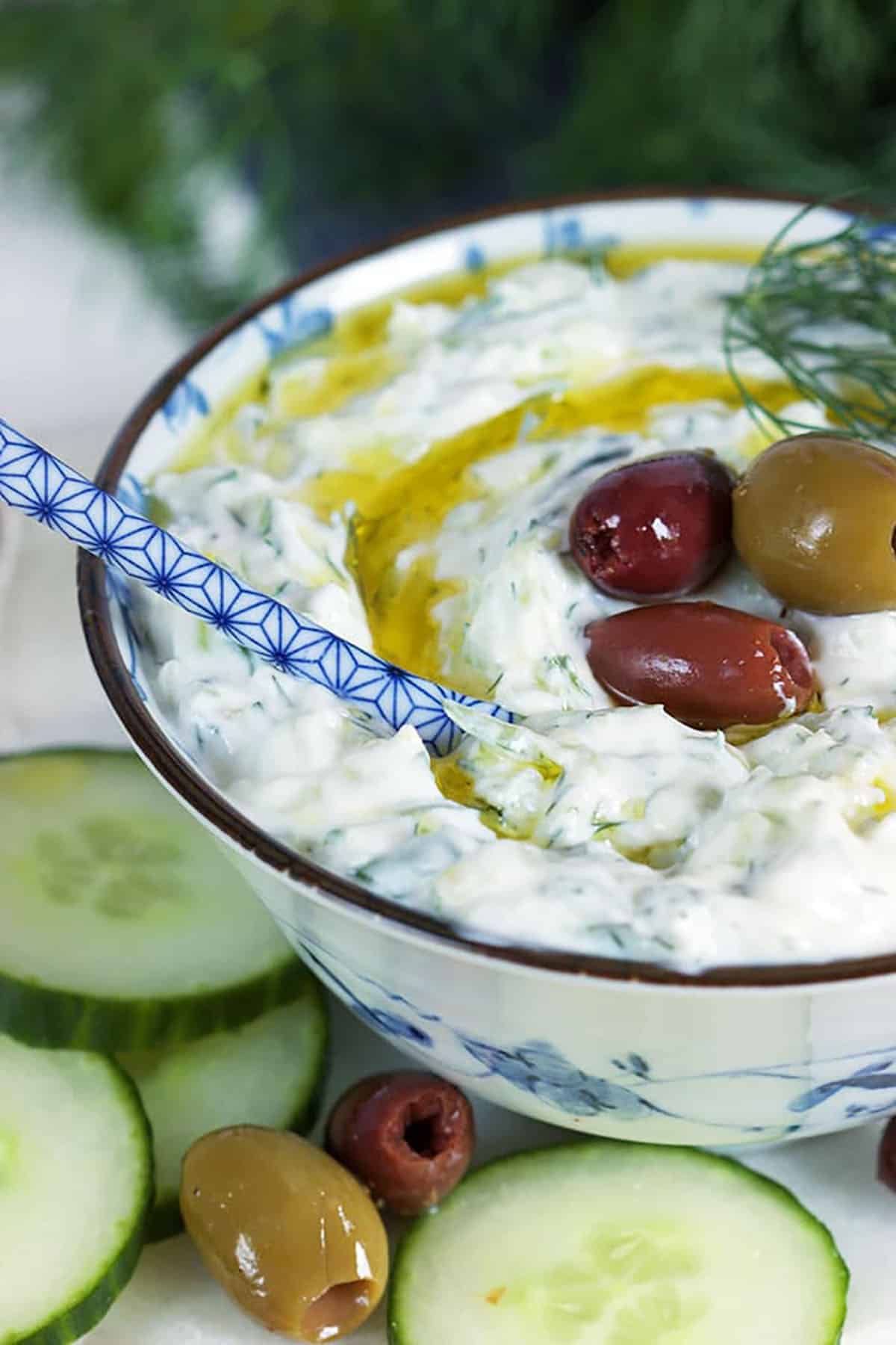 Tzatziki sauce in a white floral bowl with a blue and white spoon dipped into it and topped with dill, olives and olive oil.