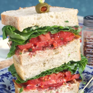 Two halves of BLT sandwich stacked on a blue and white plate with a toothpick and olive on top.