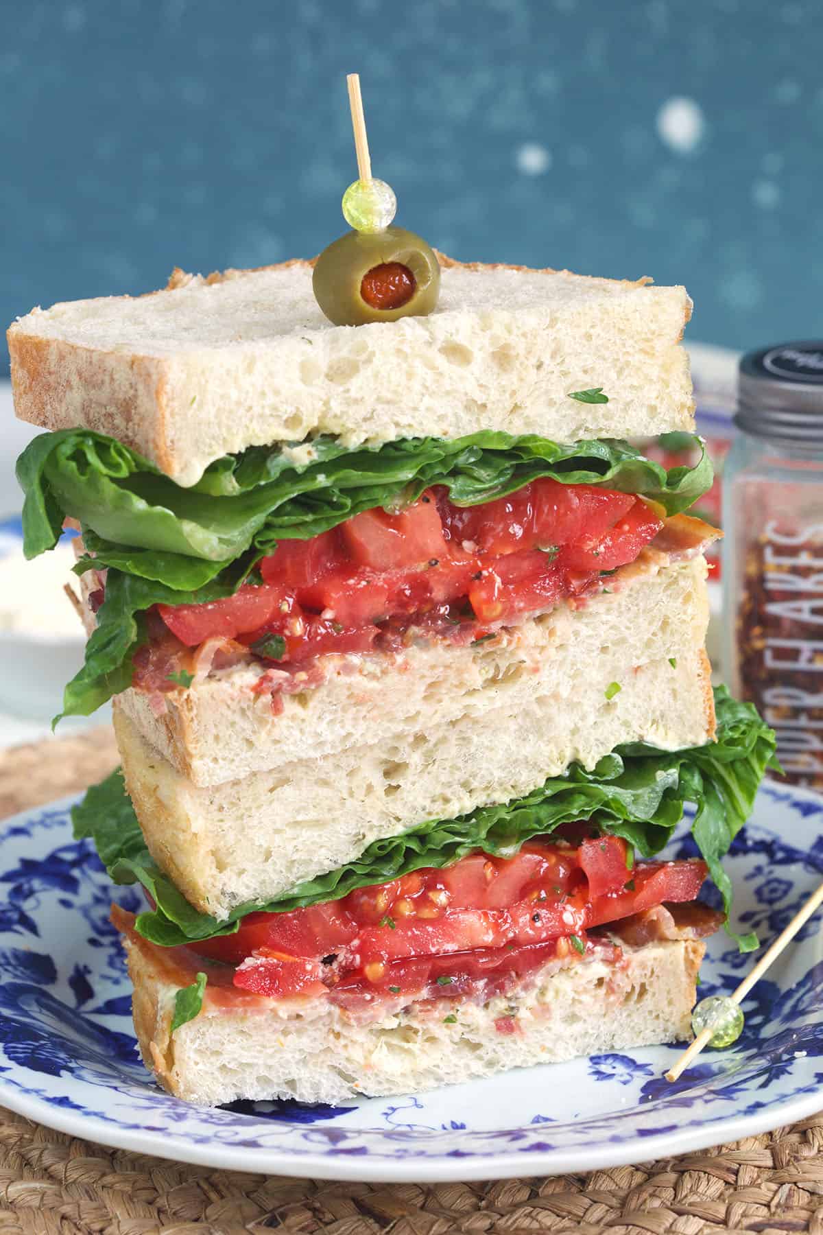 Two halves of BLT sandwich stacked on a blue and white plate with a toothpick and olive on top.