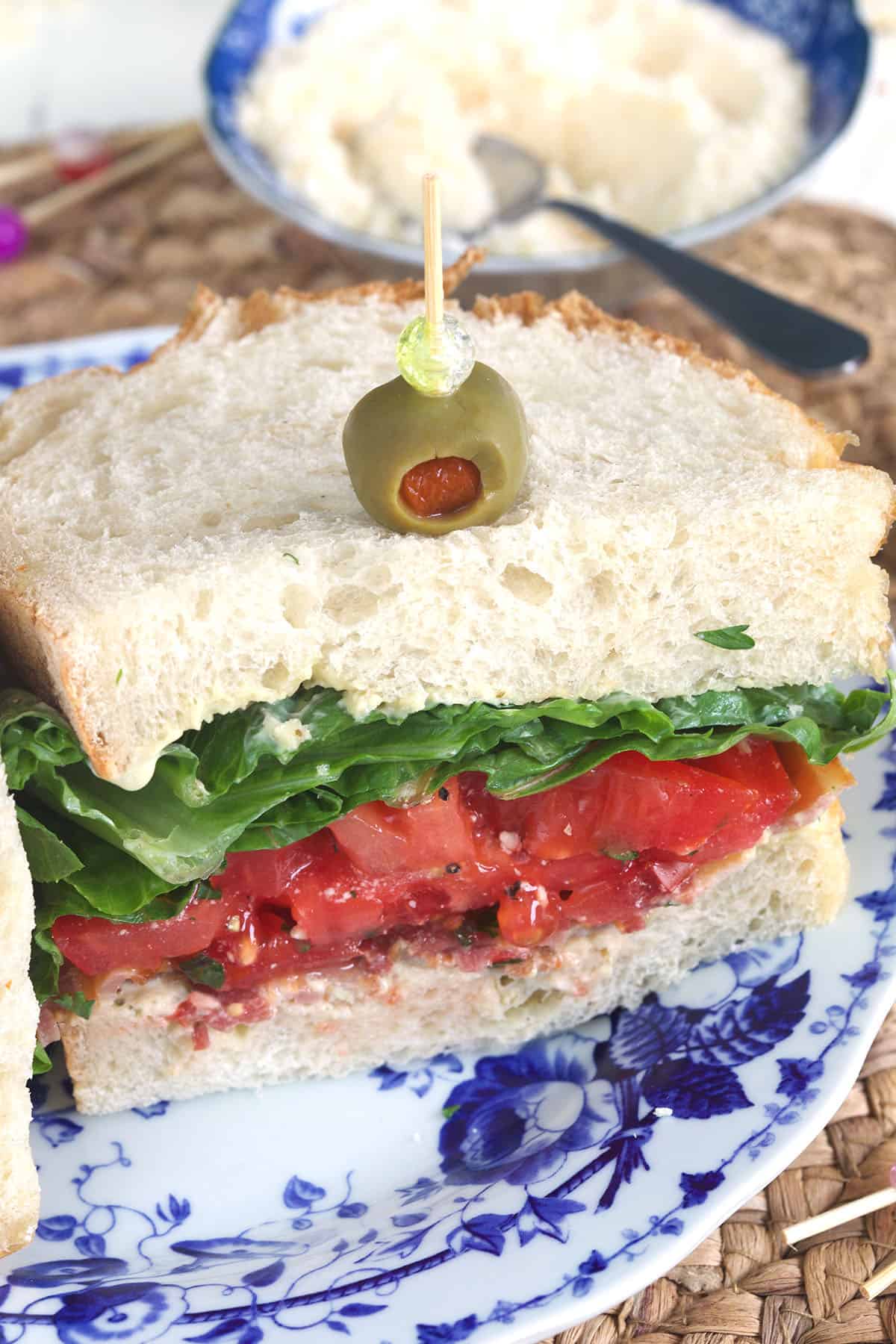 Half a BLT sandwich on a blue and white plate. A toothpick with an olive is inserted into the top of the sandwich.