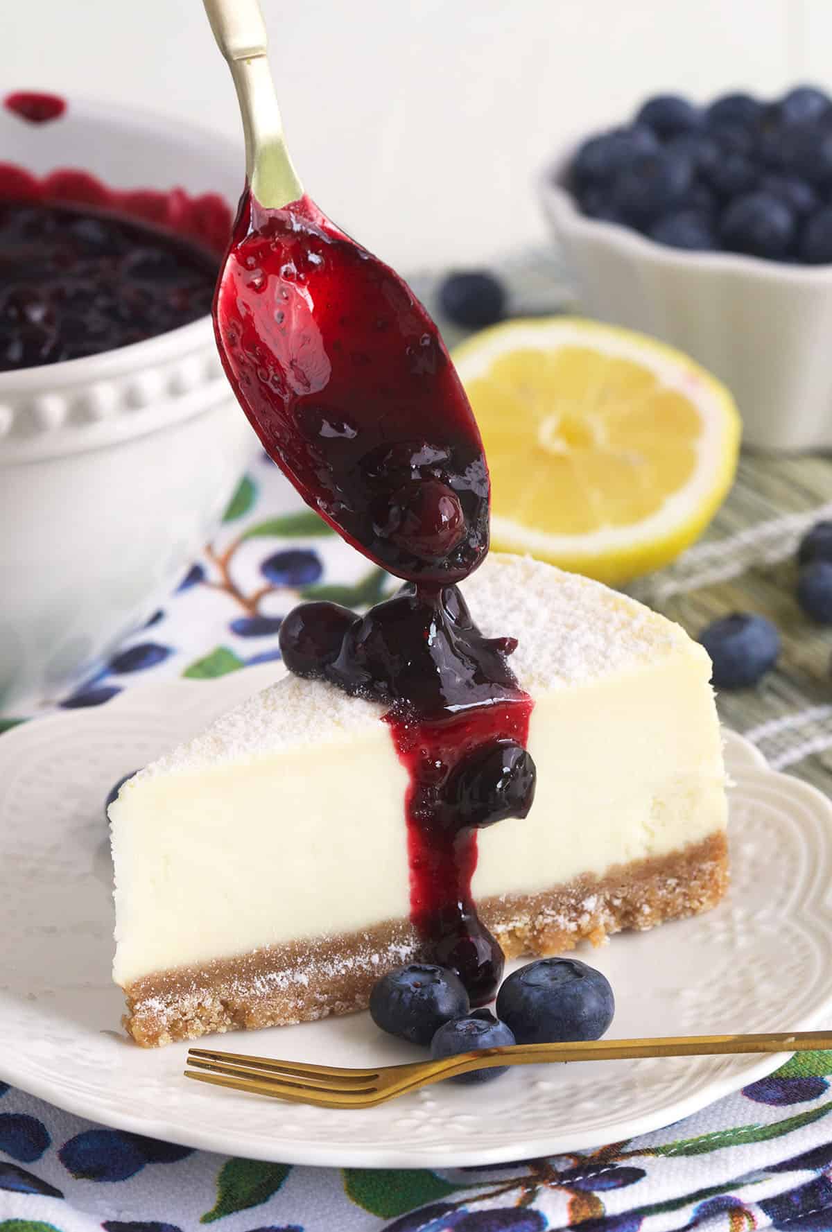 Blueberry compote is being drizzled over a slice of cheesecake. 
