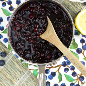 A wooden spoon is mixing blueberry compote in a silver pot.