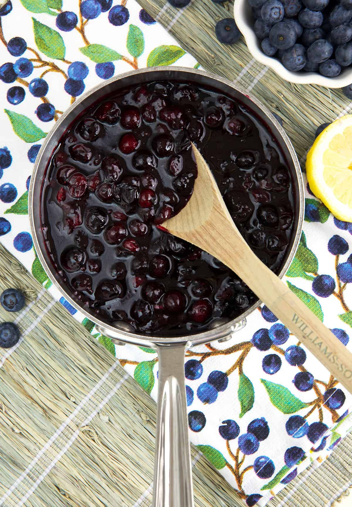 A wooden spoon is mixing blueberry compote in a silver pot. 