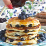 Syrup is being drizzled on top of a stack of blueberry pancakes.