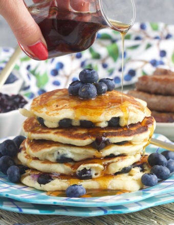 Syrup is being drizzled on top of a stack of blueberry pancakes.