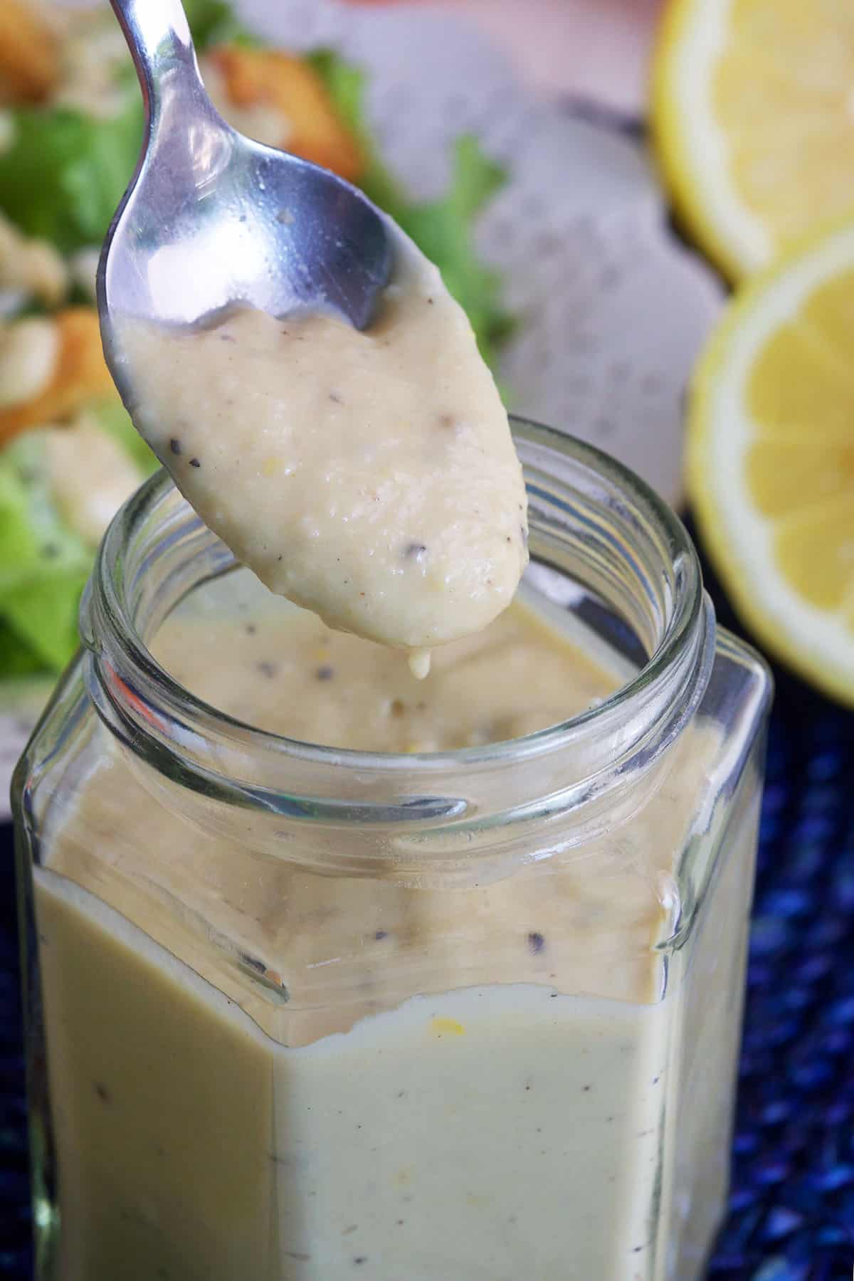 A spoon is drizzling Caesar dressing over a small glass.